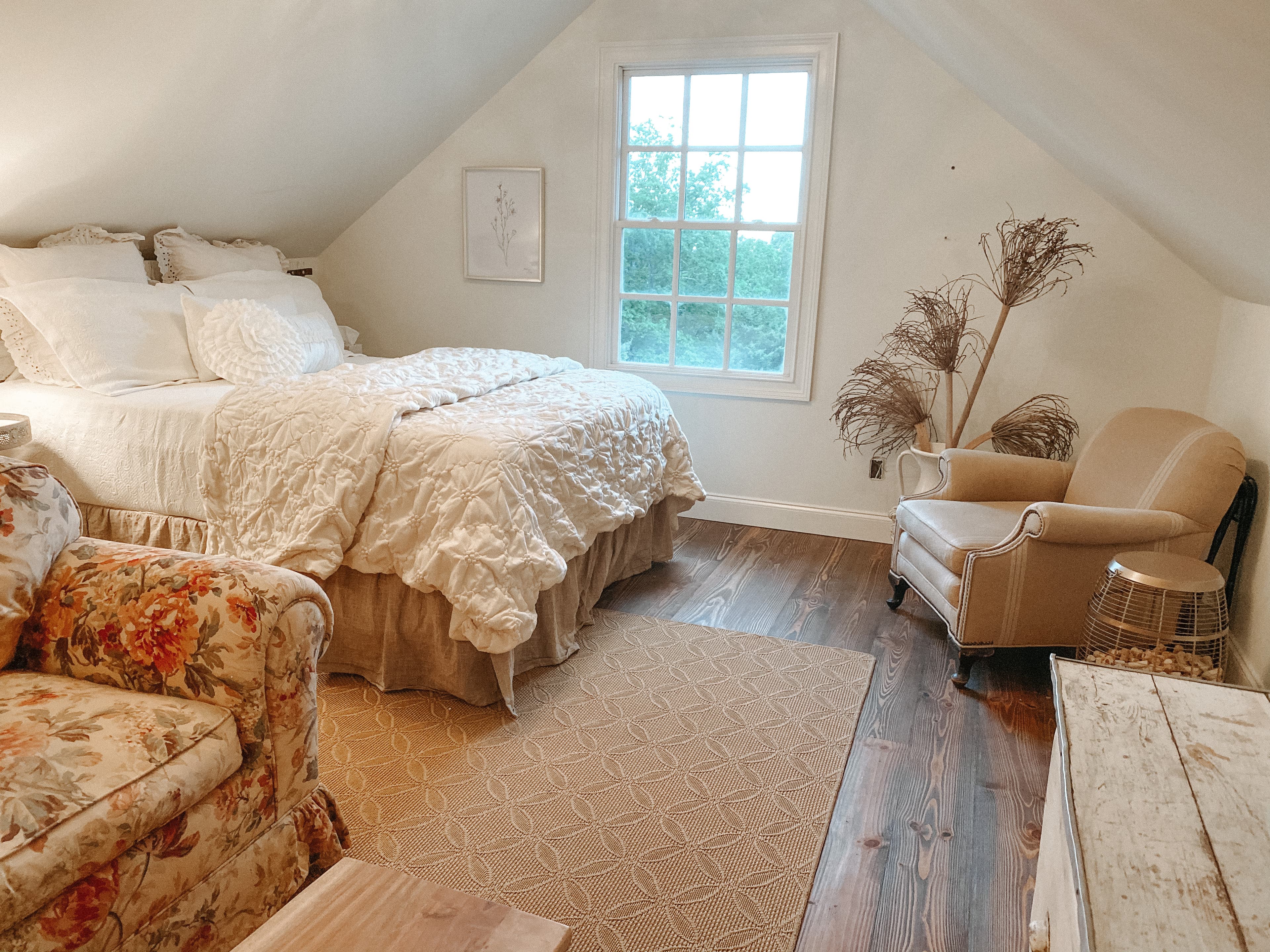 A cozy attic bedroom featuring sloped ceilings, rustic hardwood floors, a white plush bed, and vintage-style armchairs with a view of greenery through a centered window.