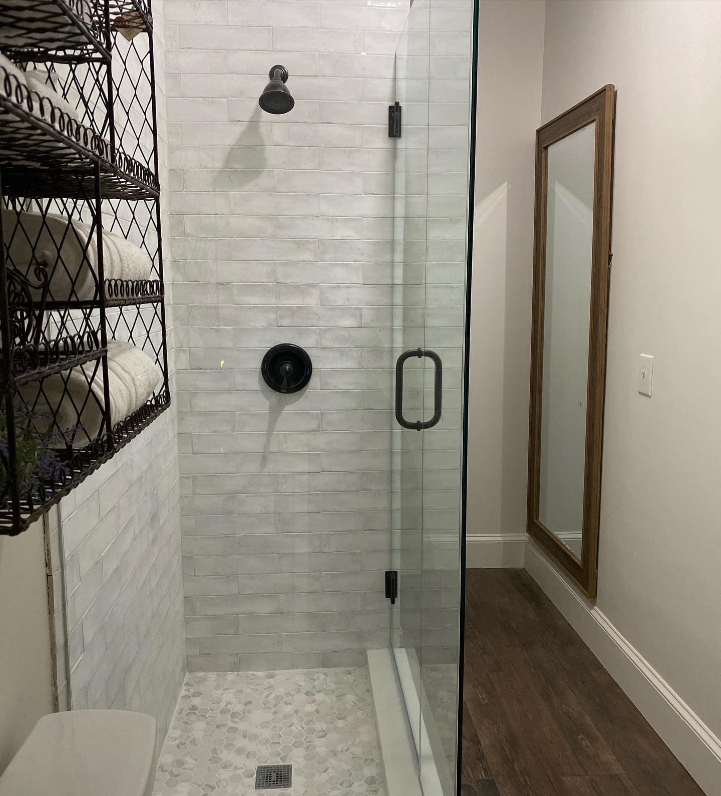 A modern walk-in shower featuring a glass door, white subway tile walls, a pebble-tiled floor, and sleek black hardware, situated next to a wire storage rack filled with white towels.