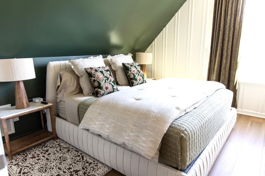 A cozy bedroom featuring a plush white bed with floral-patterned pillows against a deep green accent wall. The room is set in an attic with a sloped ceiling and includes rustic wooden nightstands and a large window with gold curtains.
