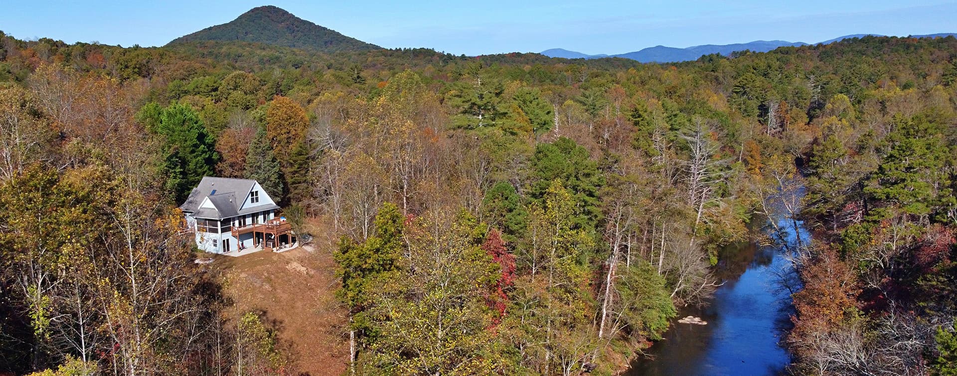 A picturesque house nestled among autumn-colored trees beside a winding river, with mountains in the distance.